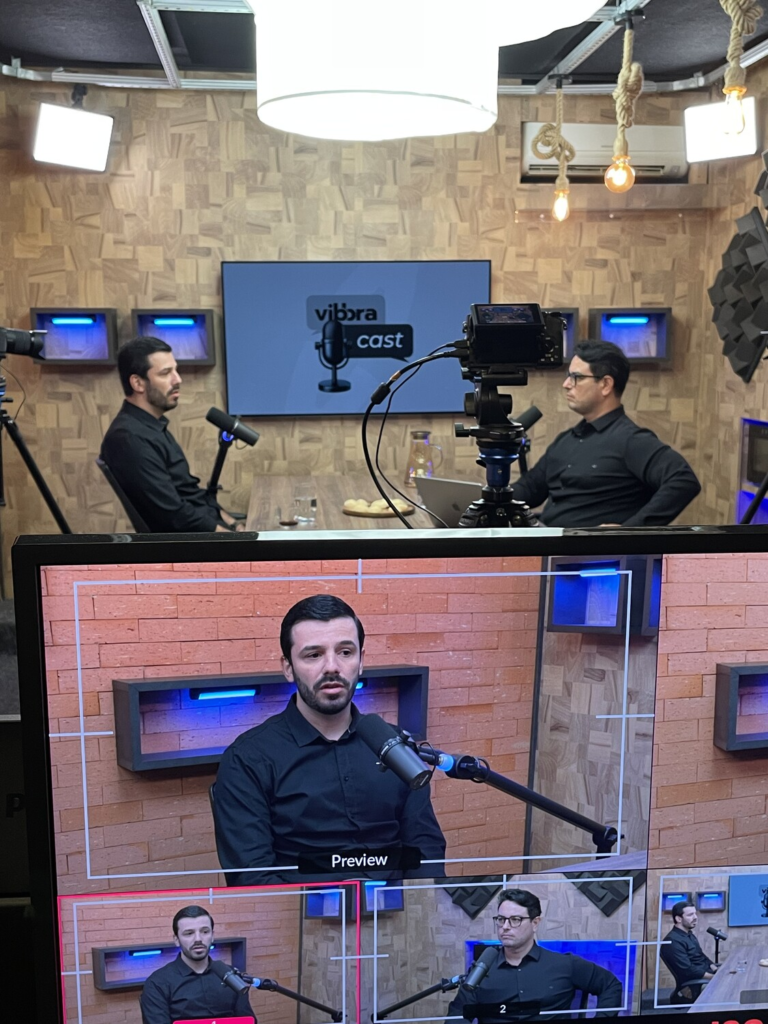 Bastidores da gravação do Vibbracast, com Leandro Oliveira e Everton Bernardes em estúdio e monitor exibindo a prévia das câmeras do podcast.
