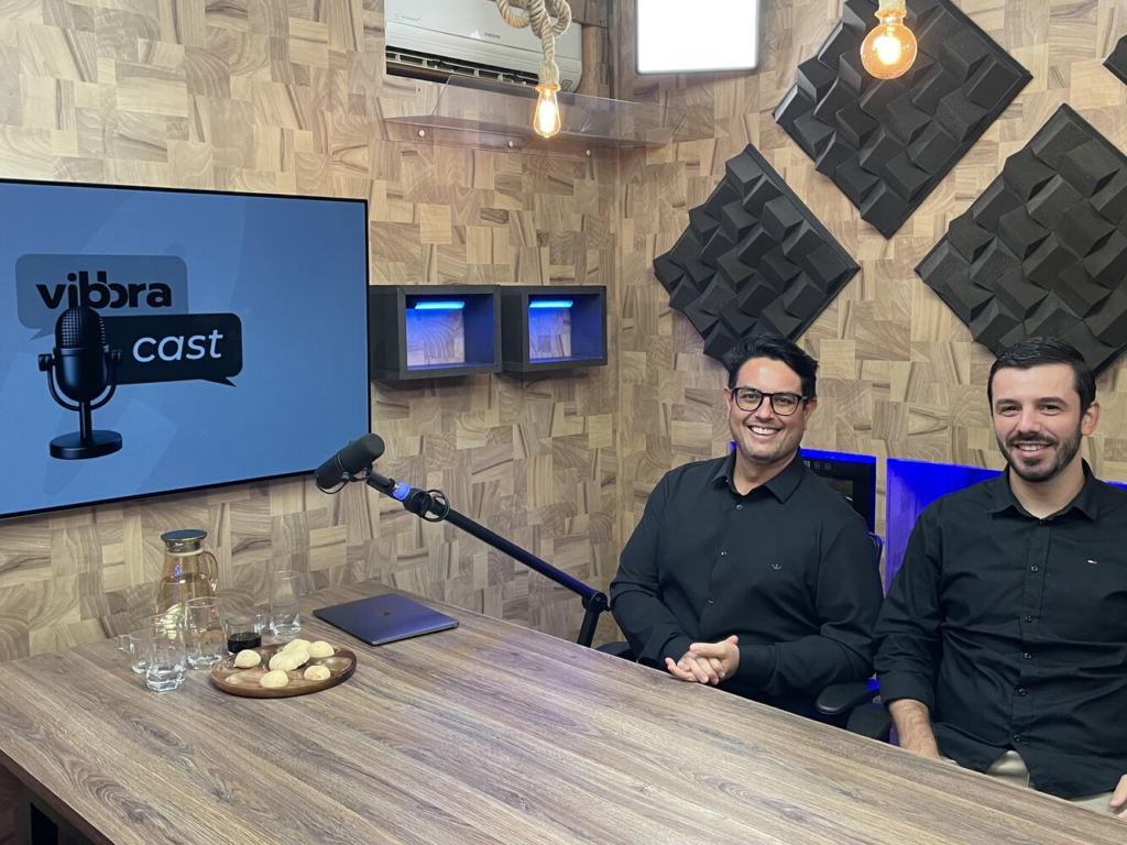 Leandro Oliveira e Everton Bernardes sorriem em estúdio de podcast da Vibbra, sentados à mesa com microfone, tela do Vibbracast e elementos de acústica ao fundo.