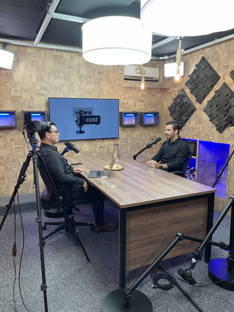 Leandro Oliveira e Everton Bernardes gravam um episódio do Vibbracast em estúdio de podcast, sentados à mesa com microfones, câmeras e iluminação profissional.