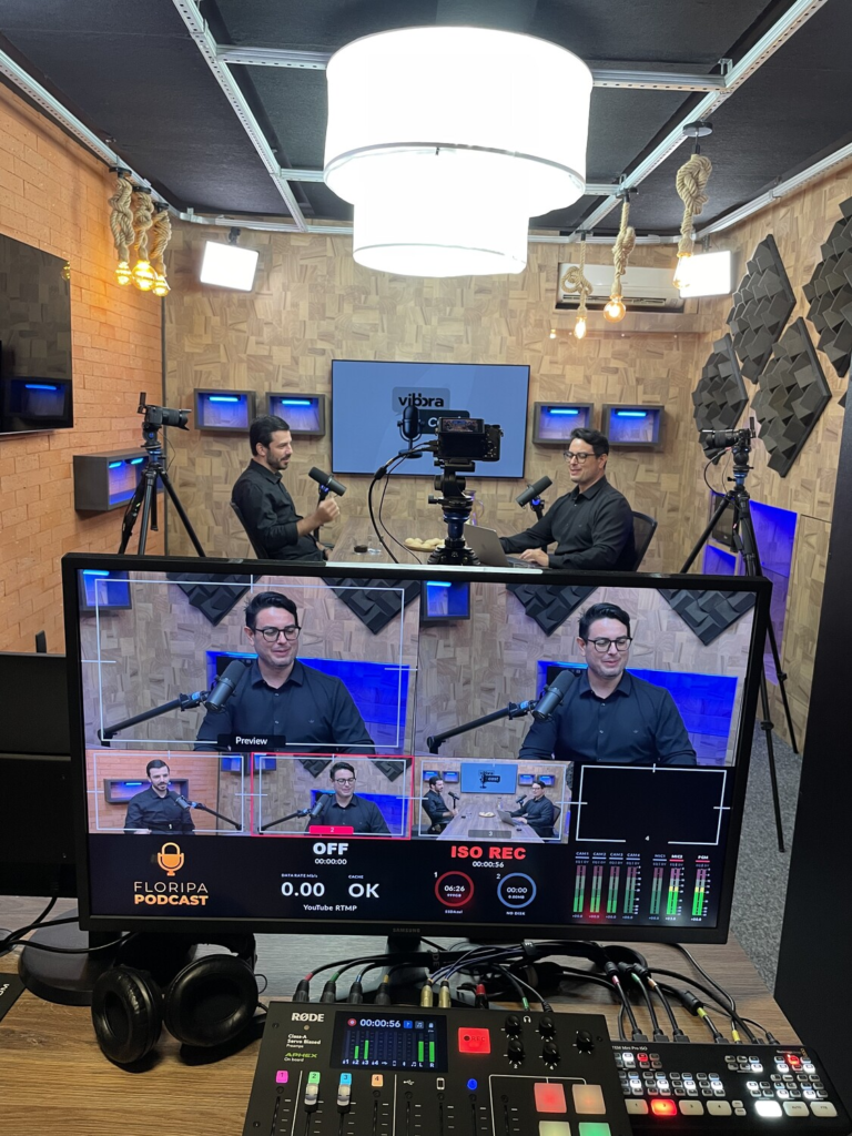 Gravação de episódio do Vibbracast em estúdio de podcast, com Leandro Oliveira e Everton Bernardes sentados à mesa, microfones, câmeras e equipamentos de transmissão.