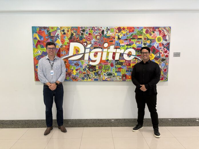 Agenor Pacheco, Diretor de Tecnologia da Digitro, e o Leandro Oliveira posando em foto na frente do logo da Digitro.