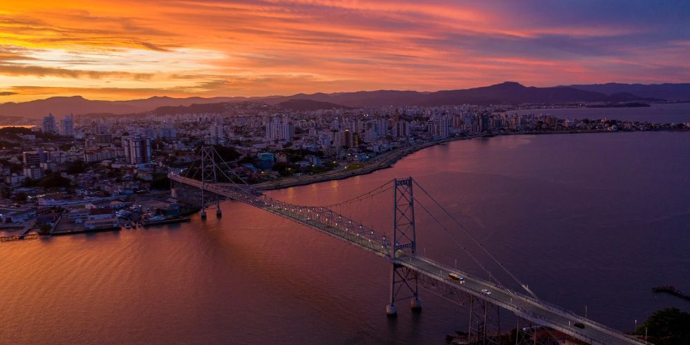Vista aérea da Ponte Hercílio Luz, em Florianópolis, ao pôr do sol, com a cidade e o mar ao fundo.
